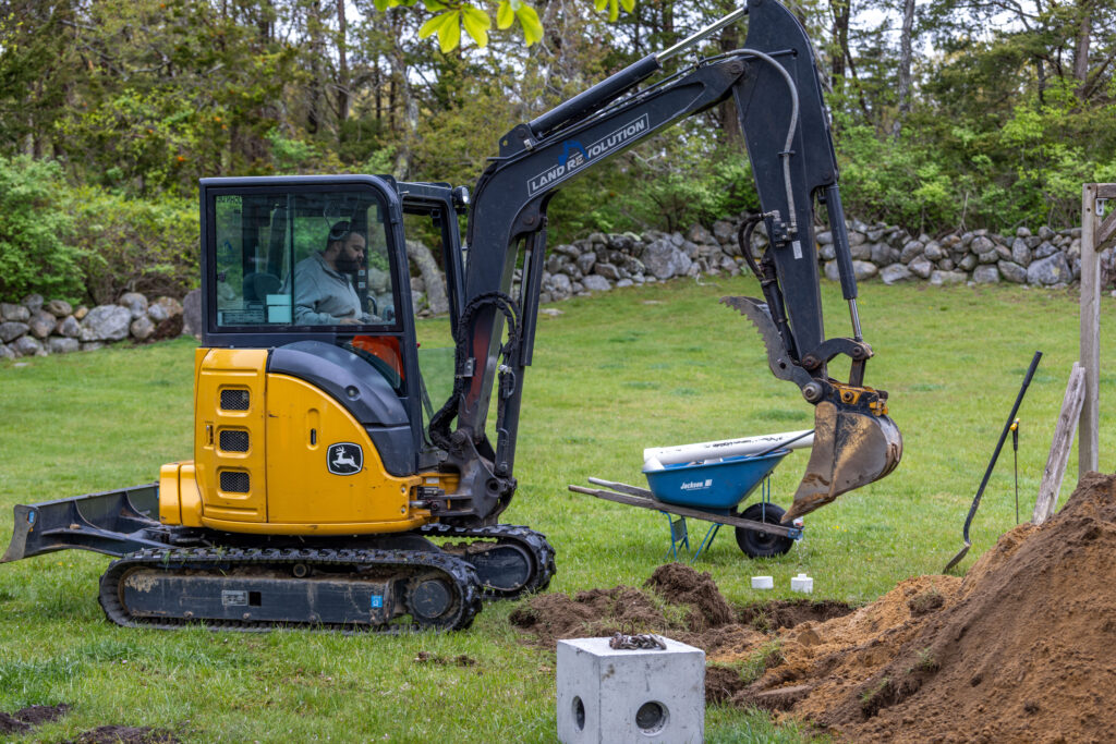 366A6038 - Land Revolution Yellow John Deere mini excavator operating on a grassy yard with dirt piles nearby and a blue wheelbarrow nearby.