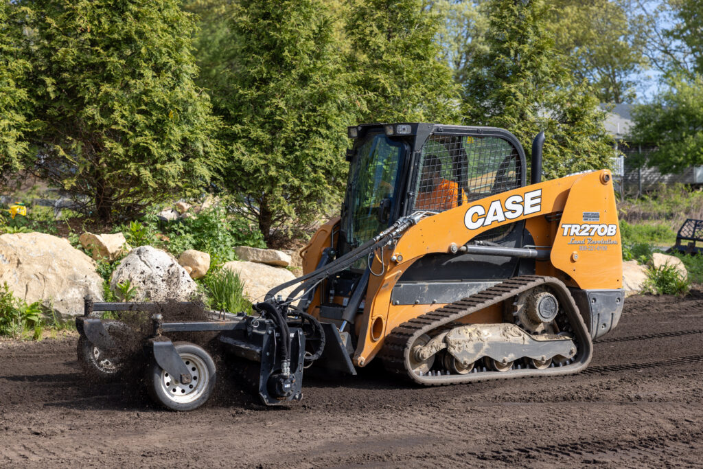 366A6168 - Land Revolution Orange CASE skid-steer loader on a dirt construction site with rocks and green hedges in the background, ready for work.