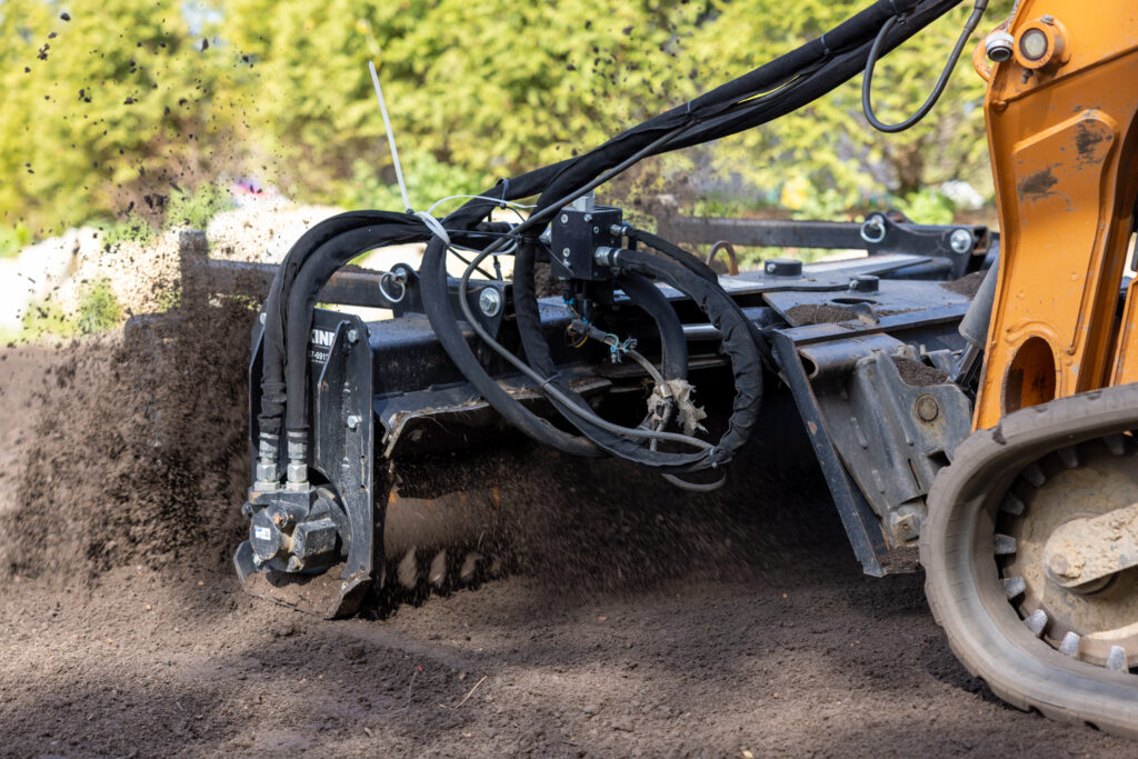366A6177 - Land Revolution Construction excavator with hydraulic arms dumping dark soil, dirt spraying into a pile at a worksite, with greenery in the background