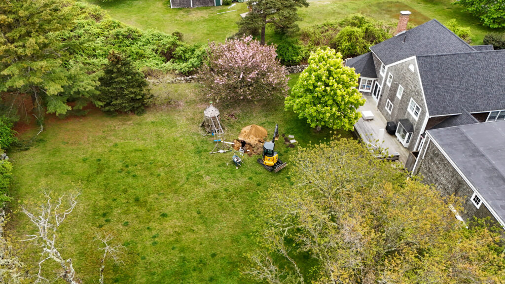 default - Land Revolution Aerial view of a backyard construction scene with a small wooden play tower, a yellow mini excavator, and a dirt mound beside a gray-shingled house.