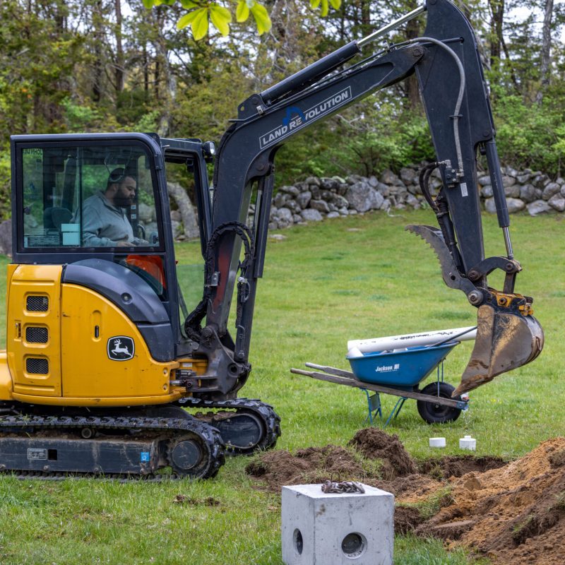 Yellow John Deere mini excavator operating on a grassy yard with dirt piles nearby and a blue wheelbarrow nearby.