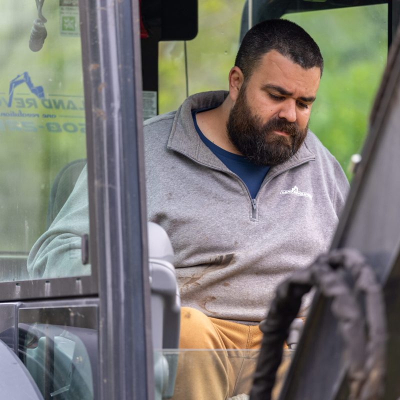 Bearded man in a gray pullover seated in a construction vehicle cab, looking down at his lap.