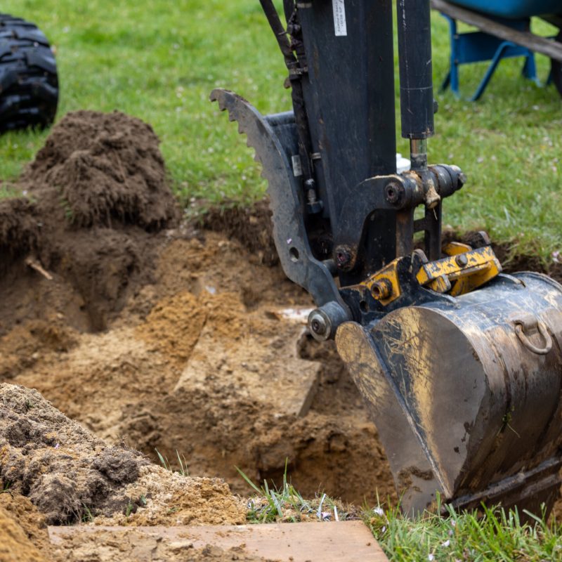 Excavator bucket digging into a dirt trench on a grassy construction site, with soil piles nearby and a worker’s leg visible in the background.