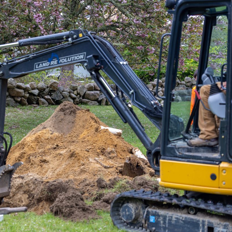 Yellow excavator with its arm digging a dirt mound in a residential garden, worker visible in the cab as it operates.