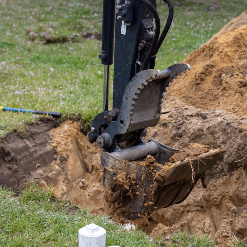 Excavator bucket with teeth digging a trench, dirt pile to the right and grassy area in the background