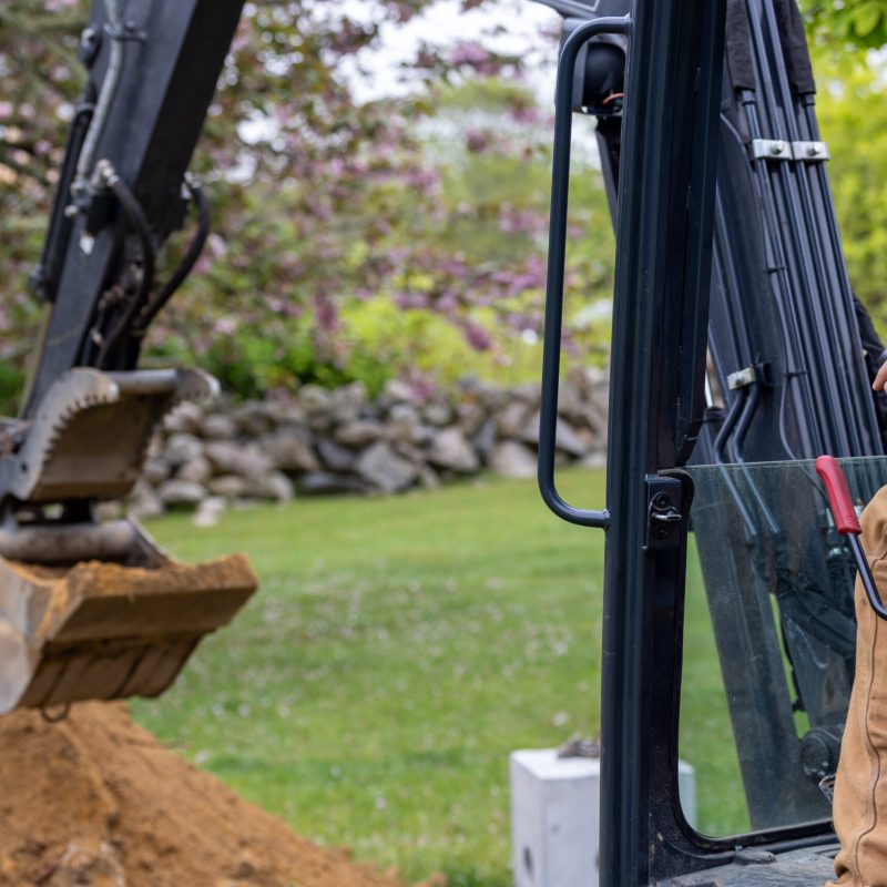 Excavator bucket dumping a load of dirt in a yard with greenery in the background, operator's hand visible in cab.