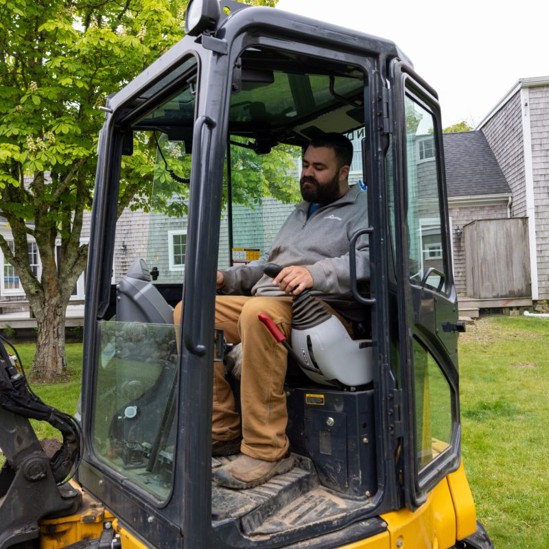 Man operating a yellow compact excavator from inside the cab in a residential yard, with a tree and houses in the background.