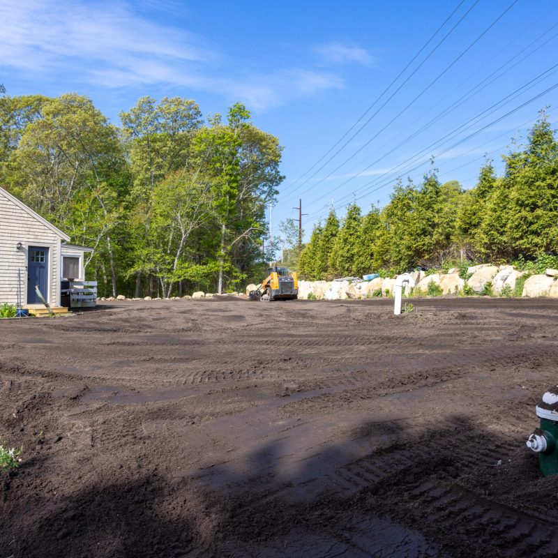 Dirt lot beside a small beige house under construction, with an excavator in the distance and trees behind, power lines across the sky.