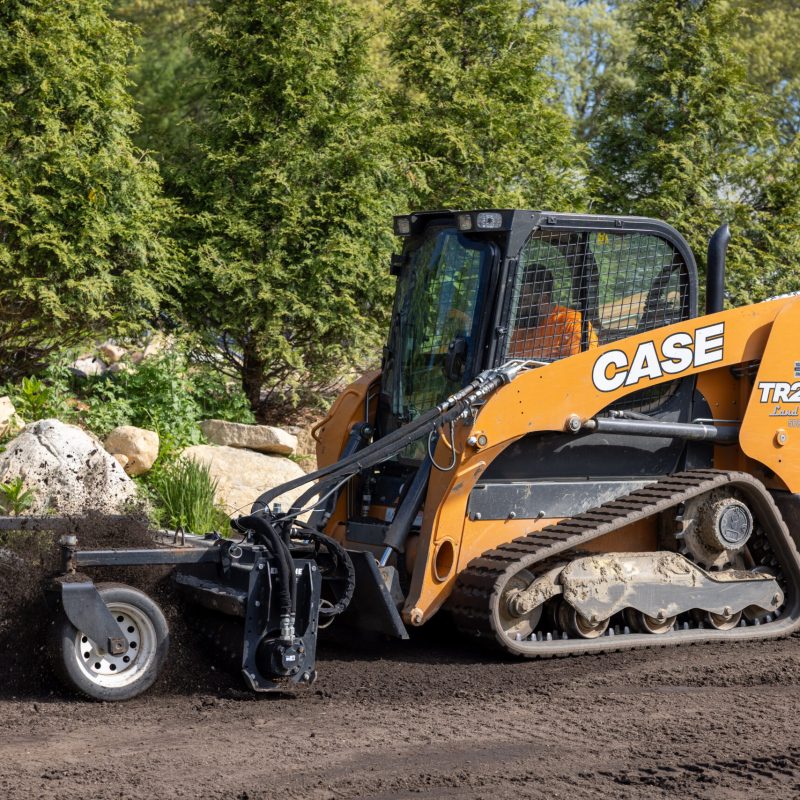 Orange CASE skid-steer loader on a dirt construction site with rocks and green hedges in the background, ready for work.