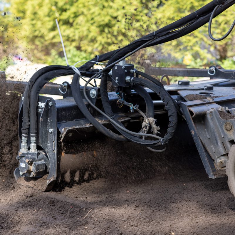 Construction excavator with hydraulic arms dumping dark soil, dirt spraying into a pile at a worksite, with greenery in the background