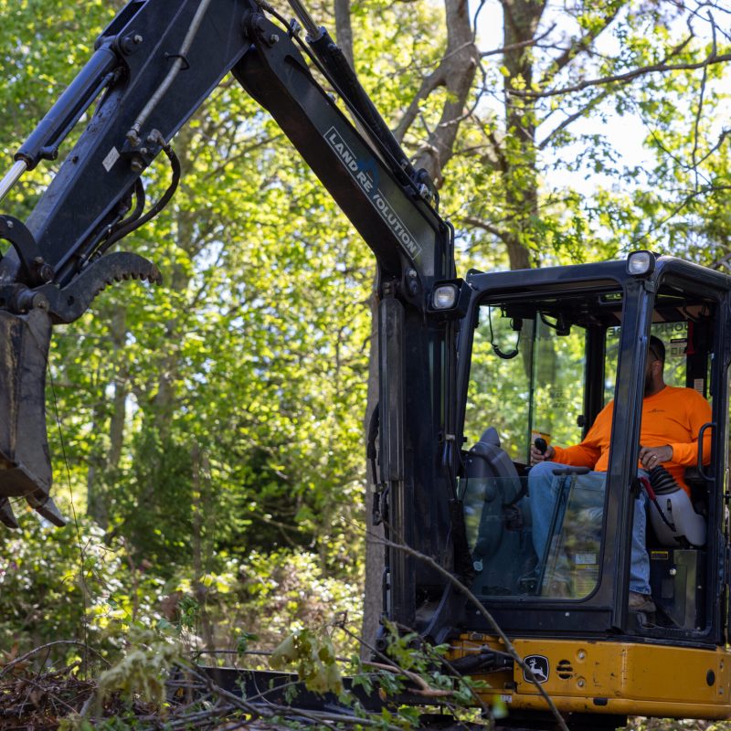 Excavator with a worker in an orange shirt operating the controls in a wooded area and green foliage around them.