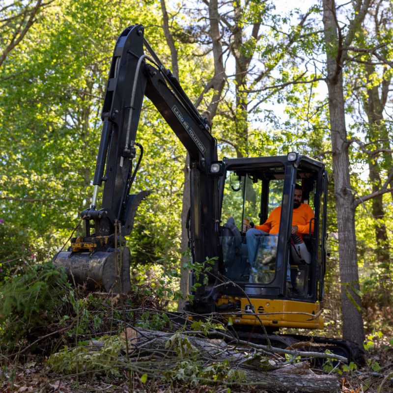 Excavator working in a wooded area with a worker in an orange shirt inside the cab, clearing brush.