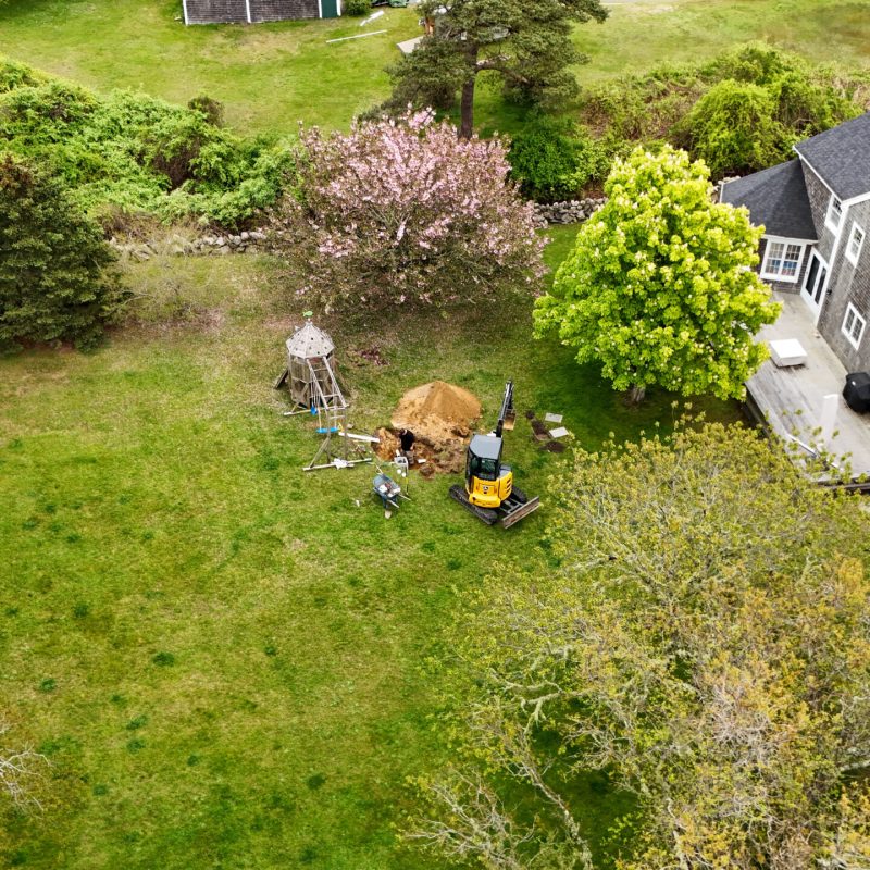 Aerial view of a backyard construction scene with a small wooden play tower, a yellow mini excavator, and a dirt mound beside a gray-shingled house.
