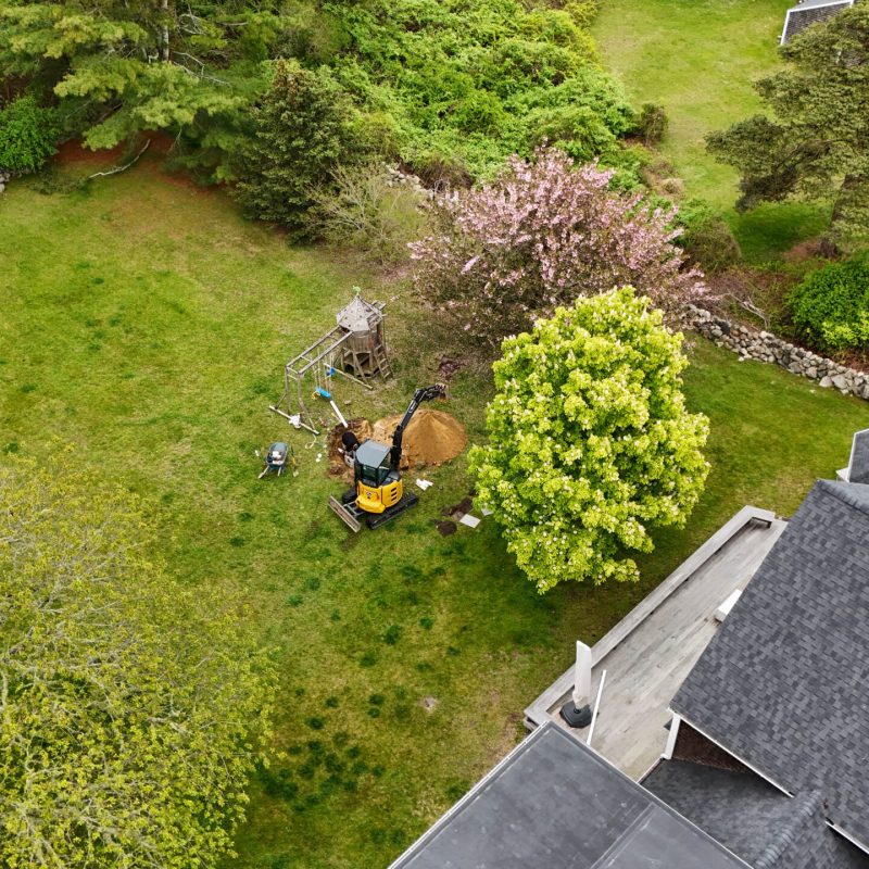 Aerial view of a backyard with a wooden playset, dirt pile, small excavator, and flowering trees beside a stone wall.