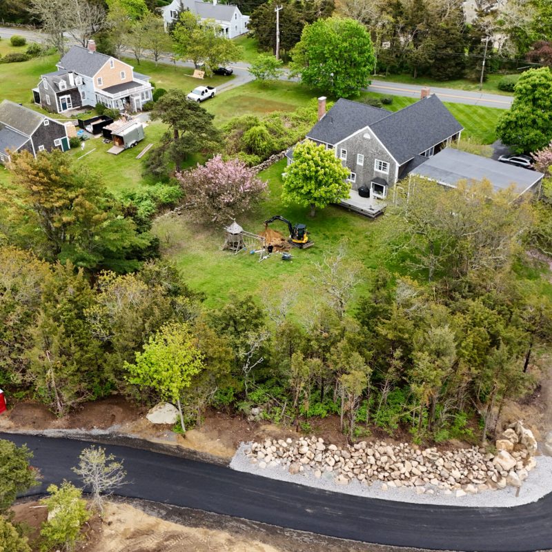 Aerial view of a suburban street and yards with a curved newly paved road, trees, and construction equipment on a lawn nearby.