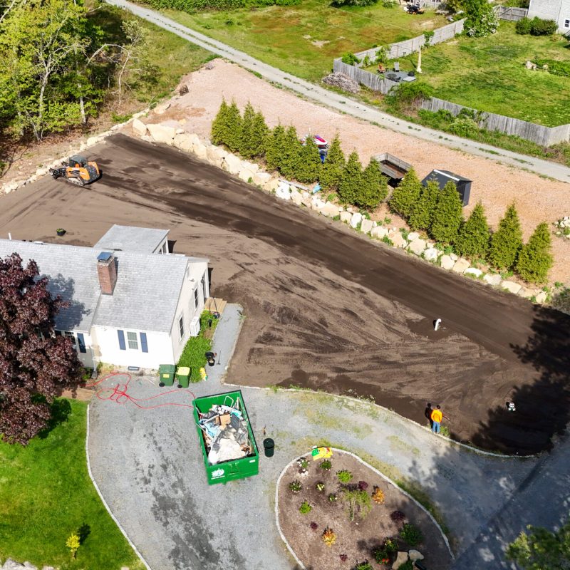 Aerial view of a residential yard under construction with a dark newly paved driveway, orange compact machine, and a green dumpster nearby.