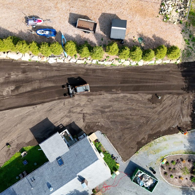 Aerial view of a construction site with a bulldozer on dark dirt, a row of green shrubs, boats on gravel, and a house with a gray roof nearby at bottom-left.