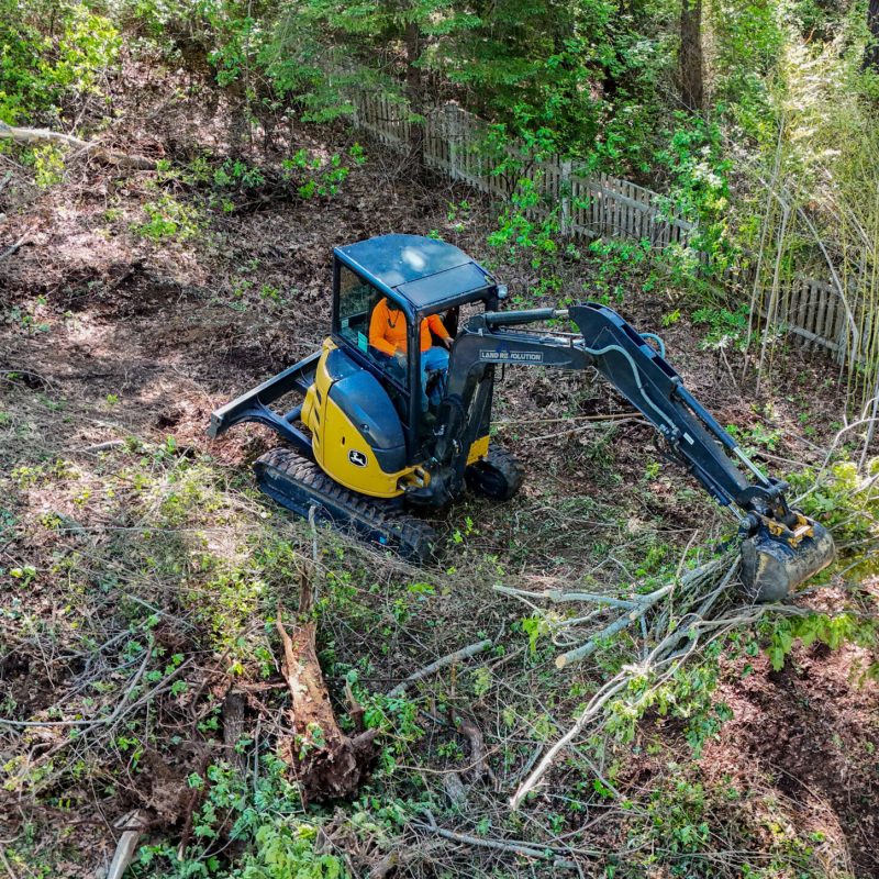 Yellow and blue John Deere mini excavator digging in a dirt trench in a wooded backyard, operator in orange shirt visible in cab.