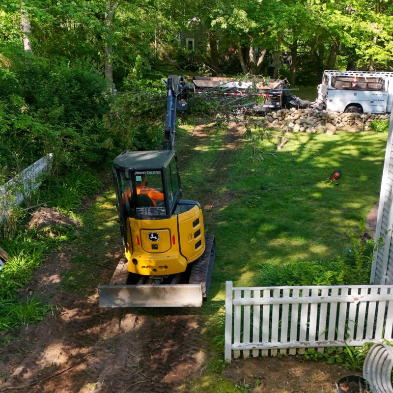 Yellow John Deere mini excavator in a backyard, moving soil while trees and a white fence surround the work area.
