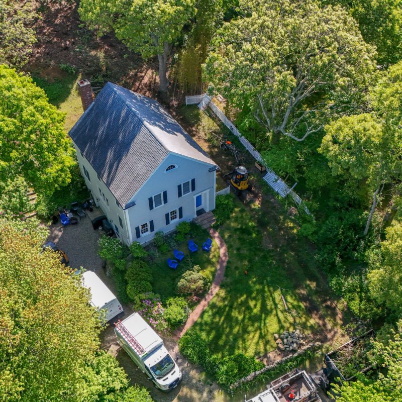 Aerial view of a blue two-story house surrounded by dense green trees; a white truck and construction equipment in the yard.
