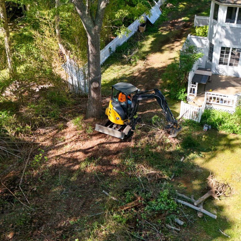 Aerial view of a backyard construction scene with a yellow mini excavator digging near a white house with a blue door.